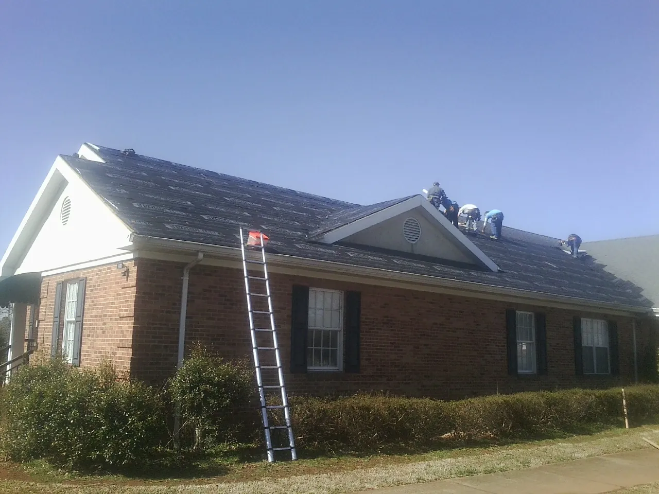 Crew tearing shingles off a brick ranch roof with a ladder set — Metal Roof Repair work by our crew in Ashby